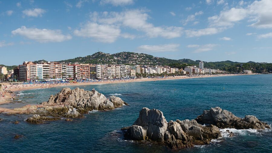 Panoramic view of Lloret de Mar, Spain, featuring a rocky beach and a distant city skyline.