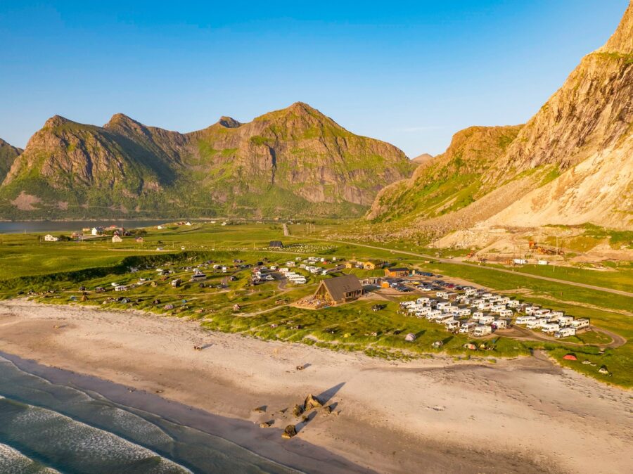 Aerial view of a Lofoten Beach Camp with rows of RVs near a sandy beach, surrounded by lush green fields and majestic mountains under a clear blue sky.