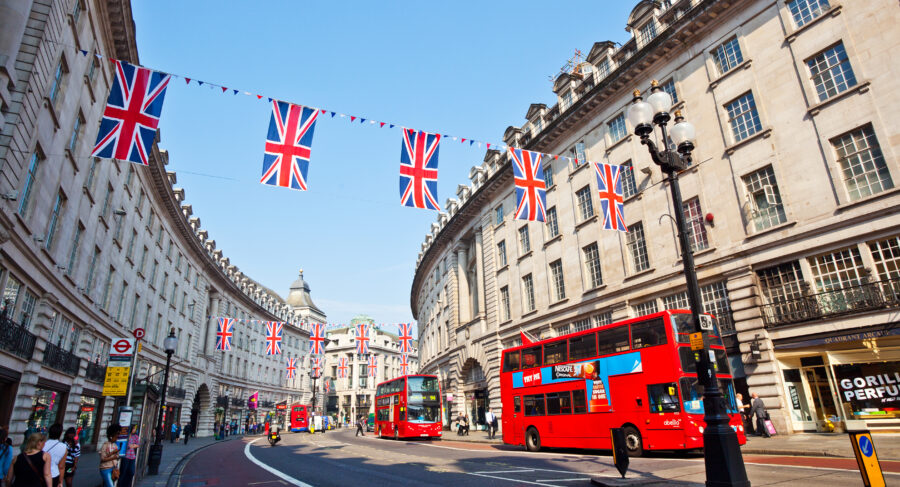 London street scene with red buses, historic architecture, Union Jack flags, and vibrant pedestrian activity.