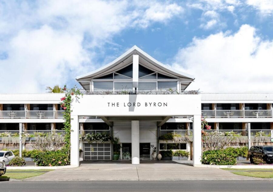 The Lord Byron Hotel front facade with balconies, portico entrance, parking, tropical landscaping, blue sky.