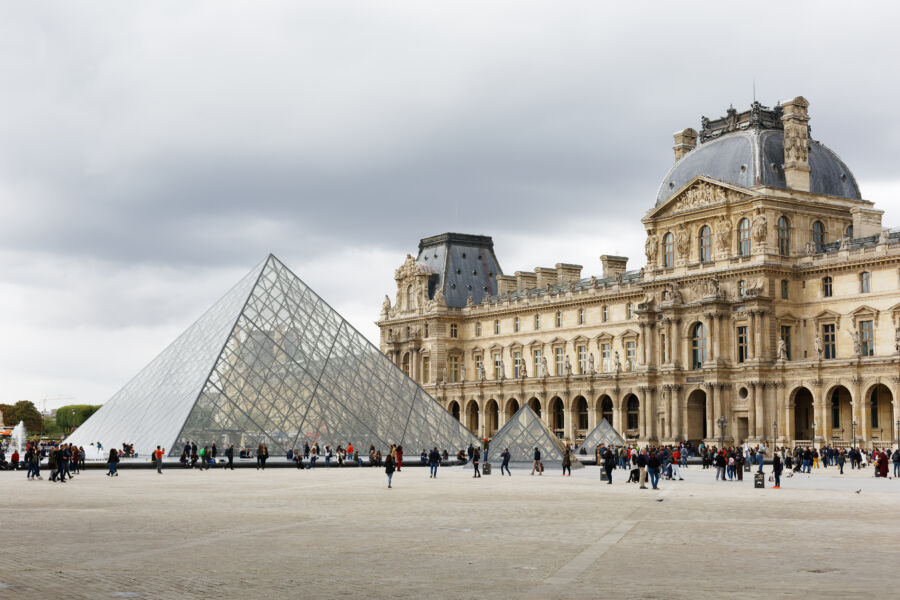 The Louvre Museum with the iconic glass Louvre Pyramid in front, set against the sky in Paris, France
