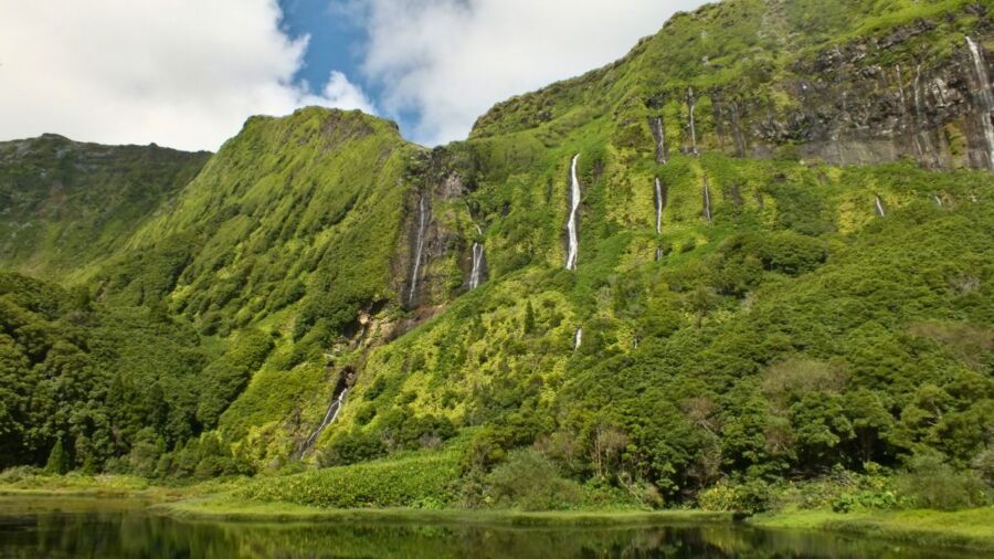 Lush green mountain with waterfalls, rocky surfaces, and reflection on calm water.
