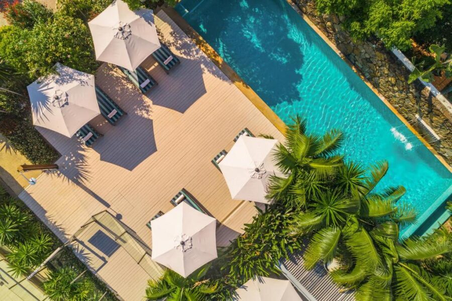 Luxury resort pool with sun loungers, umbrellas, tropical plants and wooden deck, aerial beachfront view.