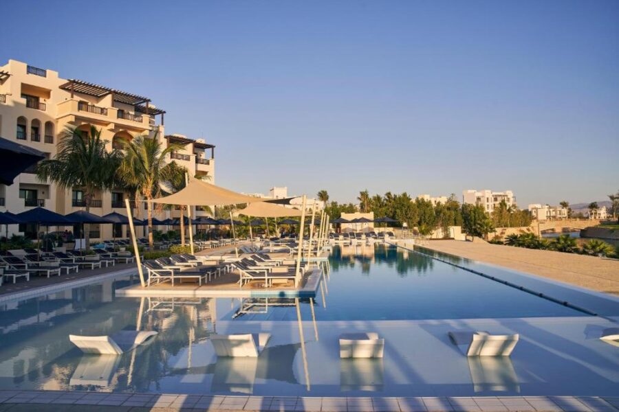 Luxury oceanfront resort infinity pool with sun loungers, palm trees, modern hotel building and clear sky.