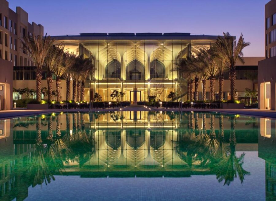 Luxury Muscat beachfront resort with illuminated glass façade, palm trees, and turquoise infinity pool at dusk.