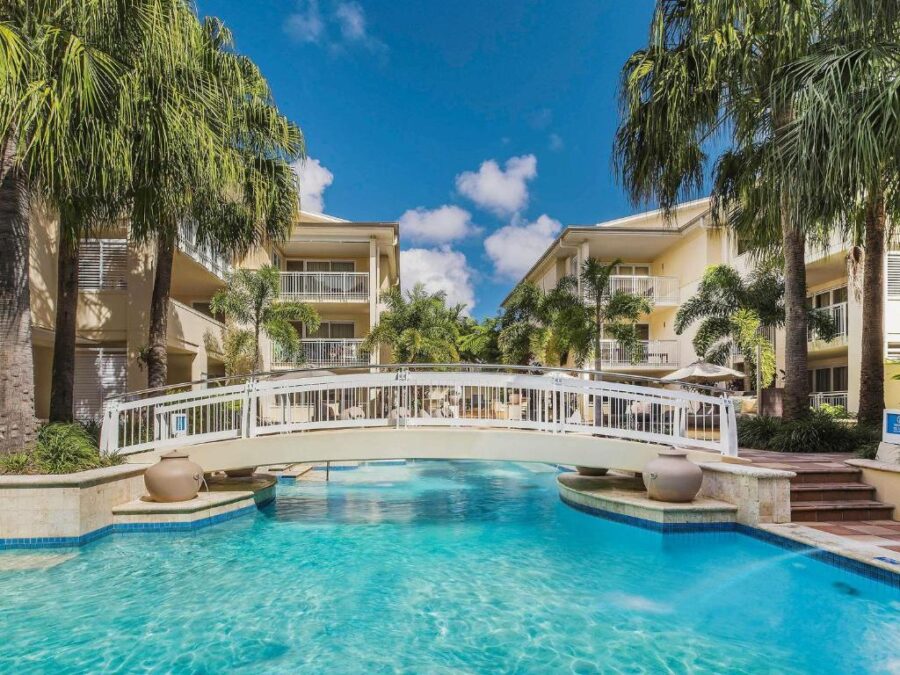 Luxury resort pool with white bridge, palm trees, lounge chairs and sunny tropical hotel buildings.