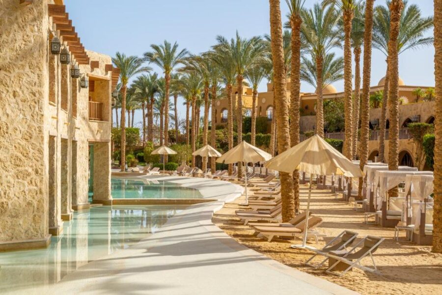 Luxury Middle Eastern beach resort pool with palm trees, sun loungers, umbrellas, and spa hotel.