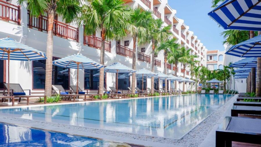 Luxury Hoi An resort with long infinity pool, palm trees, sun loungers, and modern white buildings.