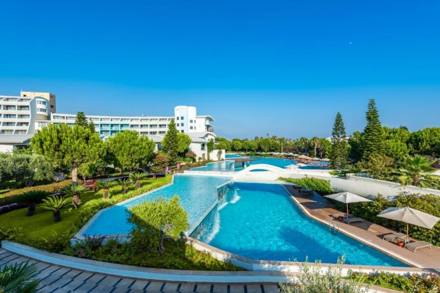 Luxury beach resort with tiered swimming pools, sun loungers, palm trees and modern hotel building.