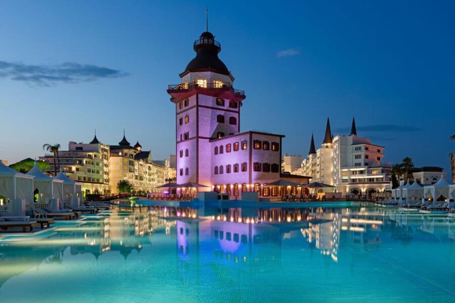 Luxury seaside resort with illuminated pool, castle-style hotel tower, and cabanas at twilight.
