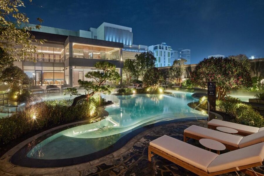 Luxury hotel rooftop pool at night with city skyline, glowing water, lounge chairs, lush landscaping.