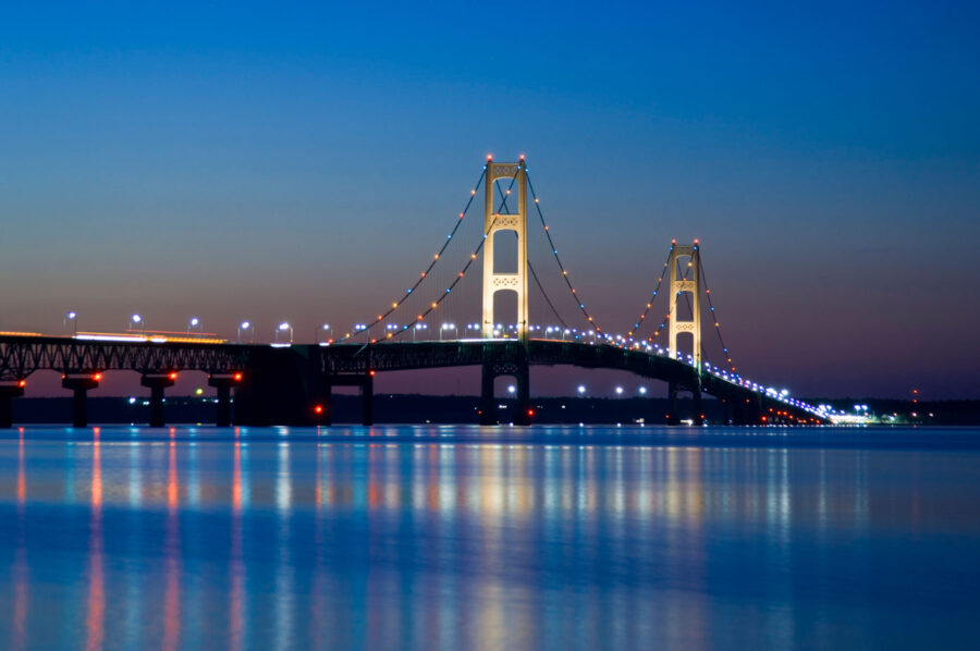 Mackinac Bridge at night with lights reflecting on calm water in summer twilight.