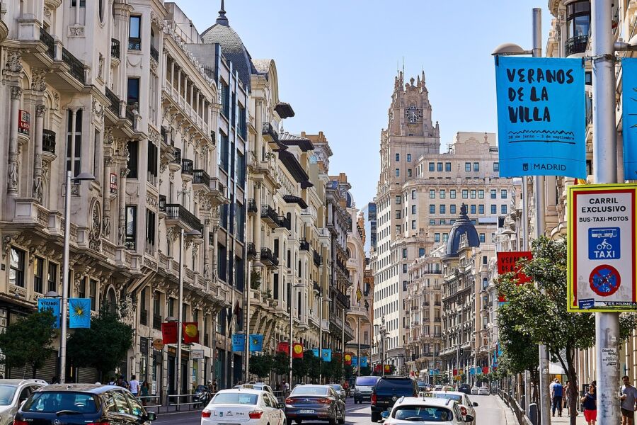 Vibrant Madrid street scene with diverse architecture, summer festival banners, and bustling city life.