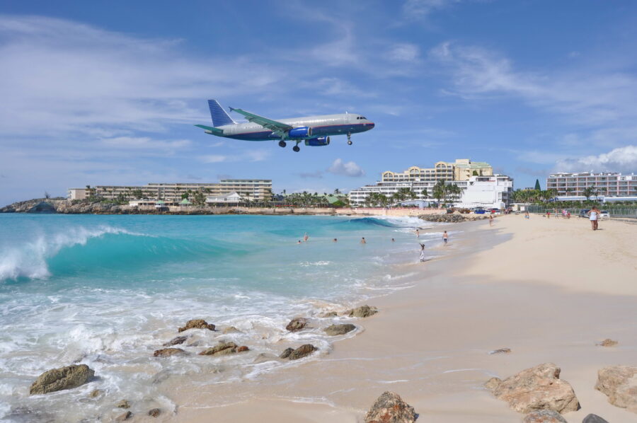 Airplane approaches landing over Maho Beach in St. Maarten, with tourists relaxing on the sandy shore