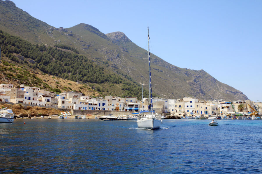 Sailboat on serene blue waters approaches a coastal village with white buildings, set against a backdrop of green hills under a clear sky in Marettimo, Sicily, Italy