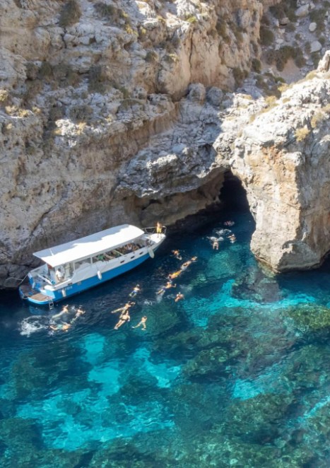 Boat with a sunshade floats beside a rocky cliff, near a small cave. Swimmers enjoy the clear, vibrant turquoise water, creating a lively summer scene.