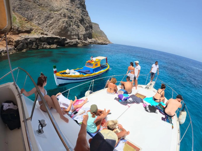People relax on a boat's deck, sunbathing and talking, as it sails near a rocky coastline. A colorful boat passes by in the clear blue water.