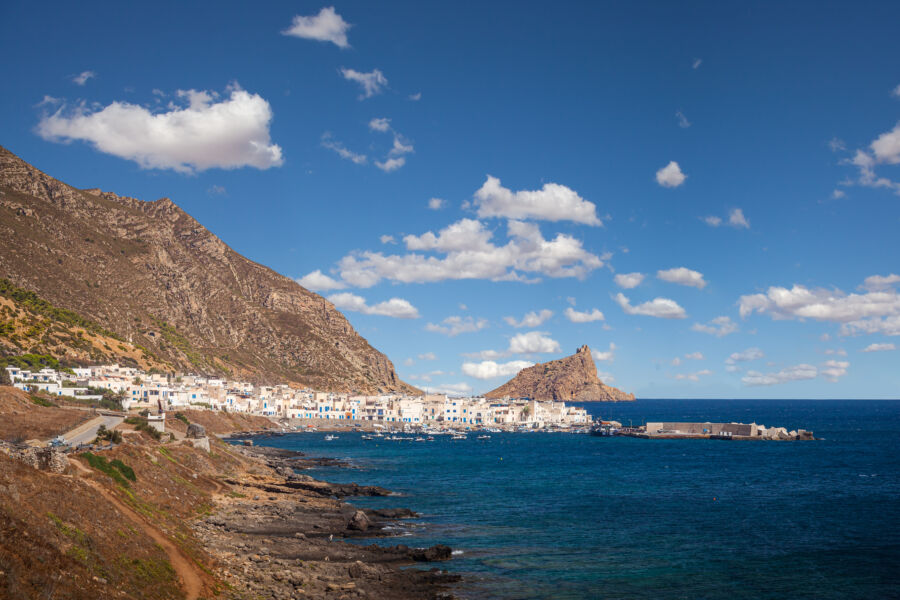Marettimo Village, Sicily, Italy with white buildings sits by a vibrant blue sea under a clear sky with scattered clouds. Rugged mountains rise in the background.