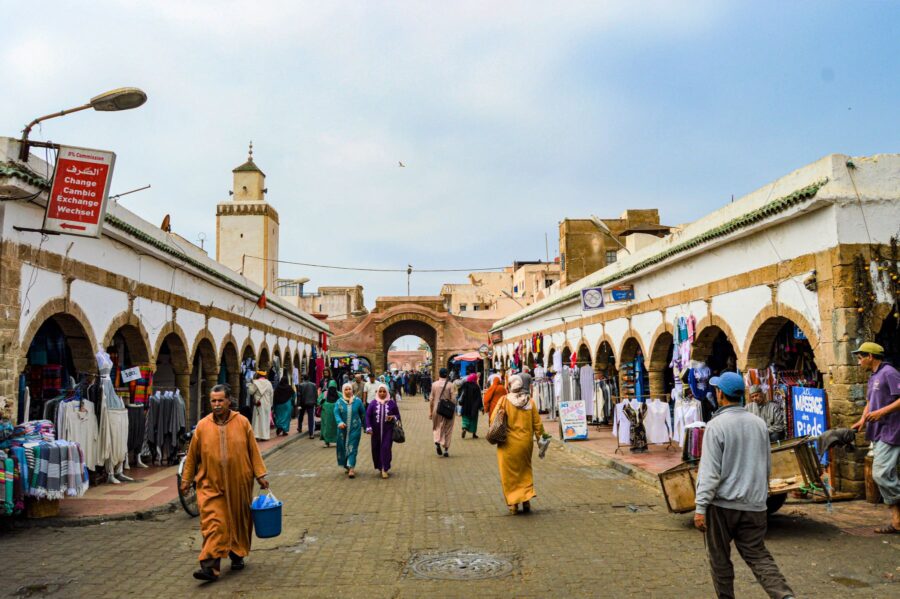 Bustling Moroccan medina market street with arcades, stalls, mosque minaret, locals shopping and walking.