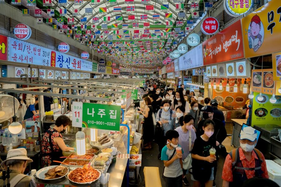 Vibrant Gwangjang Market scene with diverse food stalls, lively crowd, and multicultural ambiance.