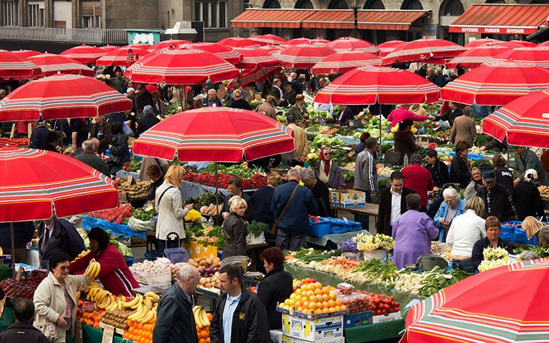 Vibrant city market scene with diverse shoppers, fresh produce, and red-striped umbrellas.