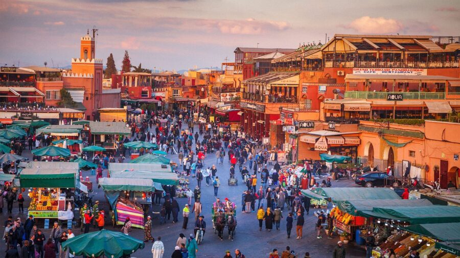 Bustling Marrakech market scene with vibrant stalls, diverse crowd, and traditional Moroccan architecture.