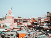 Bustling Marrakech marketplace with pink buildings, vibrant crowds, vendors, and traditional architecture.