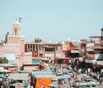 Bustling Marrakech marketplace with pink buildings, vibrant crowds, vendors, and traditional architecture.
