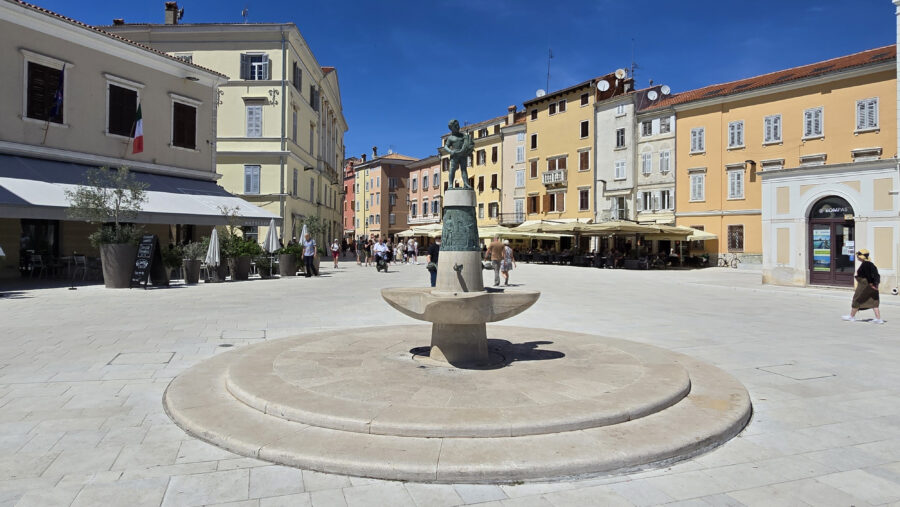 Fountain in Marshal Tito Square, Rovinj, surrounded by people walking and enjoying the lively city scene.