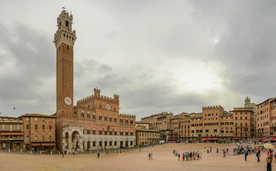 Piazza del Campo, Siena: medieval brick square with Torre del Mangia and cafes, cloudy sky.