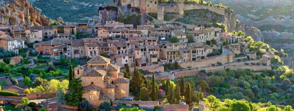 Medieval village aerial view with castle, terracotta roofs, lush greenery, and rugged landscape.