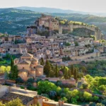 Medieval village aerial view with castle, terracotta roofs, lush greenery, and rugged landscape.