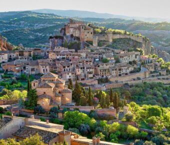 Medieval village aerial view with castle, terracotta roofs, lush greenery, and rugged landscape.