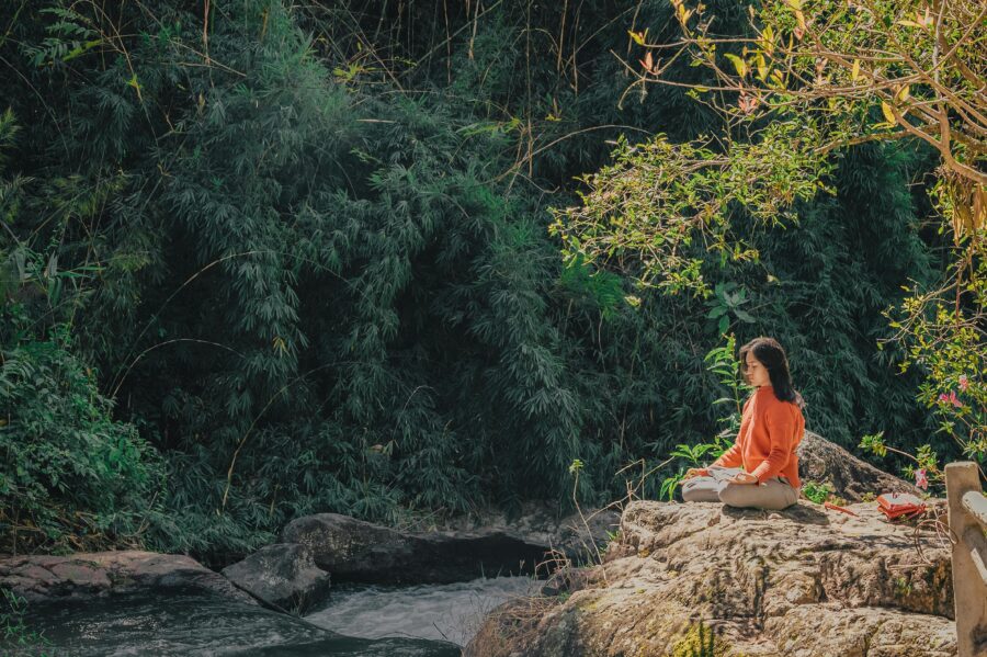 Woman meditating by a tranquil river surrounded by lush greenery and sunlight.
