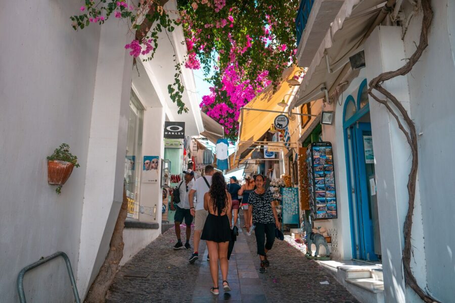 Mediterranean village alley with whitewashed buildings, cobblestones, flowering plants, and bustling shops.