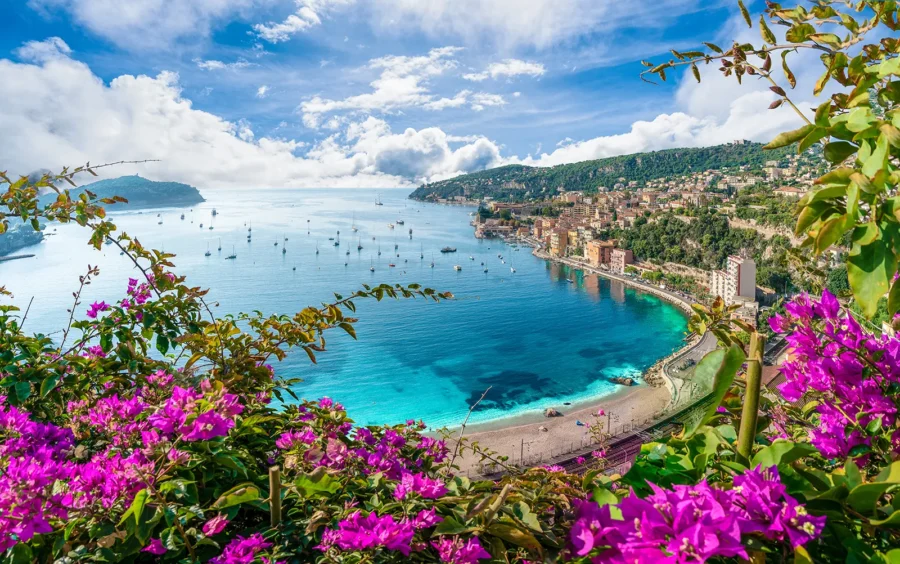 Côte d’Azur Mediterranean bay with magenta bougainvillea, sailboats, pebble beach, and hillside village.