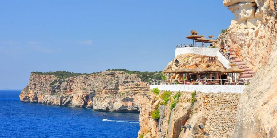 Mediterranean cliffside terraces and seaside restaurant at Caves of Xoroi overlooking deep blue sea.