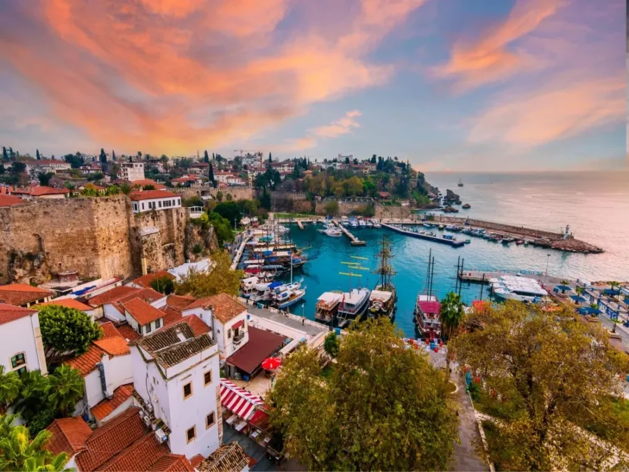 Mediterranean coastal harbor at sunset with historic stone walls, whitewashed houses, boats, and lighthouse.