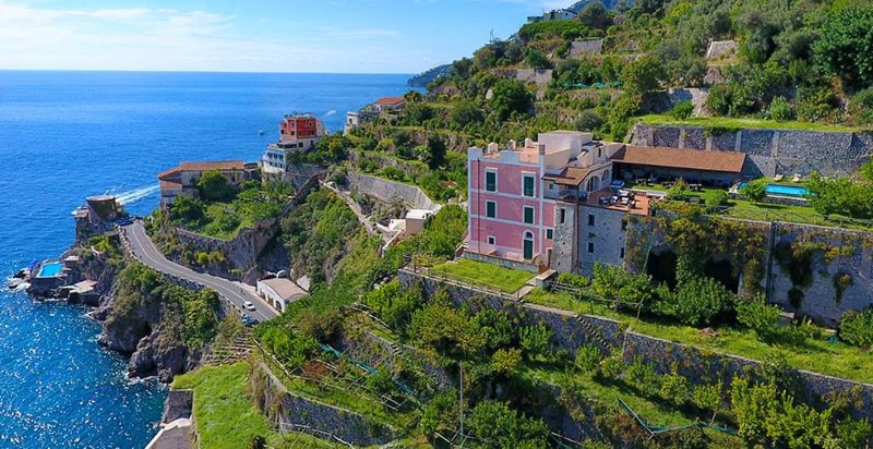 Scenic Mediterranean coastline with terraced hills, pink villa, lush greenery, and serene blue sea.