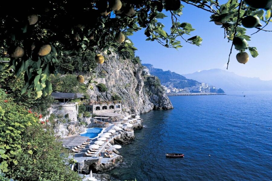 Amalfi Coast cliffs, lemon trees, beachside pool, and Mediterranean architecture under clear sky.