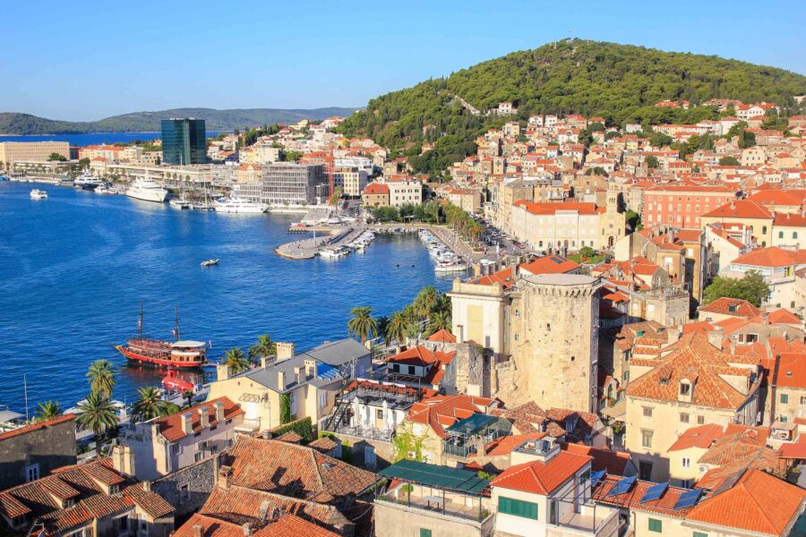 Aerial view of Mediterranean coastal town with vibrant rooftops, serene bay, and lush hills.