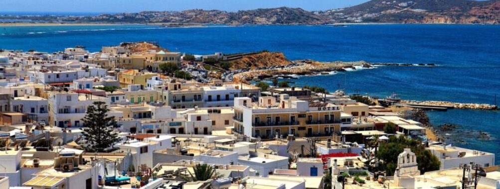 Whitewashed Greek island town with blue sea, rocky coastline, boats, hills, and serene mountains.