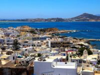 Whitewashed Greek island town with blue sea, rocky coastline, boats, hills, and serene mountains.