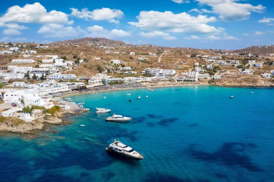 Idyllic Mediterranean coastal view with azure waters, sandy beach, yachts, white buildings, and rolling hills.