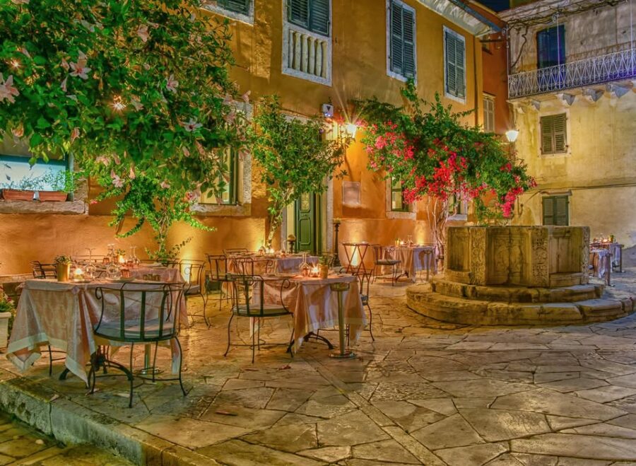 Charming Mediterranean dining scene with fountain, lanterns, floral accents, and cozy tables.