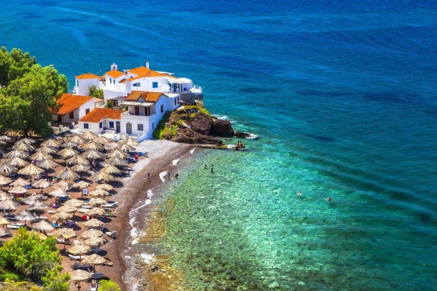 Vlichos Beach Mediterranean cove with pebble shore, turquoise water, umbrellas, and seaside village.