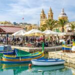 Mediterranean Malta harbor with colorful boats, bustling cafés, palm trees, and prominent church towers.