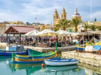 Mediterranean Malta harbor with colorful boats, bustling cafés, palm trees, and prominent church towers.