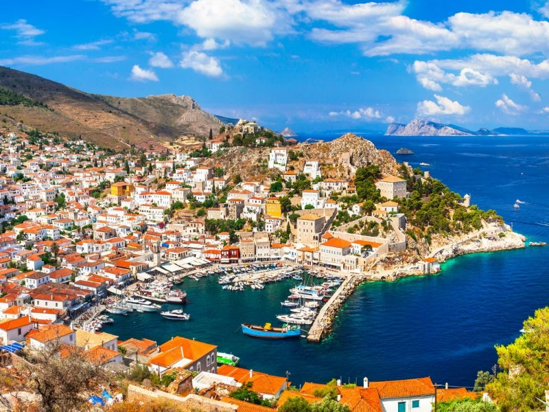 Greek island harbor with whitewashed houses, terracotta roofs, marina and turquoise sea.