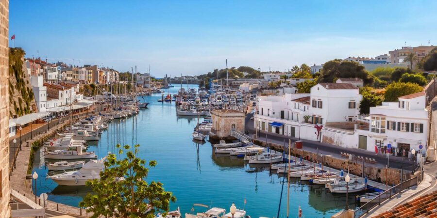 Menorca Mediterranean harbor with turquoise water, moored boats, stone quays, sunlit buildings.